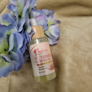 May include: A clear glass bottle with a bamboo dropper and a white label that says "Goshen Skin Science VC Turmeric Essential Oil" with the text "VC Brightening & Firming" and "Turmeric Brightening" in smaller font. The bottle is sitting on a brown fuzzy surface.