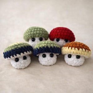 May include: Five handmade crochet mushrooms in various colors. The mushroom caps are green, red, blue, and brown, with white stems and black button eyes. The mushrooms are arranged closely together, showcasing the handmade details.