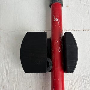 May include: A black plastic tool holder with two curved arms that hold a red pole in place. The holder is mounted on a white wall.