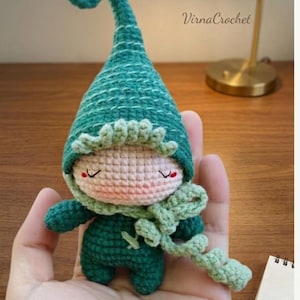 May include: A handmade crochet doll in shades of green, featuring a pointed hat with a curled tip, a scarf, and closed eyes. The doll is held in a hand. The text "Virna Crochet" is visible in the background.
