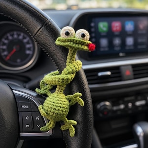 May include: A green crocheted frog figure with large white eyes, black pupils, and a small red tongue. The frog has a long, coiled body wrapped around the black leather steering wheel of a vehicle, featuring a round head and four limbs.