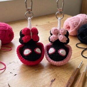 May include: Two handmade crochet keychains, one red and black, the other pink and black, each with a silver keyring. The keychains are shaped like a character with a bow. Yarn and crochet hooks are in the background.
