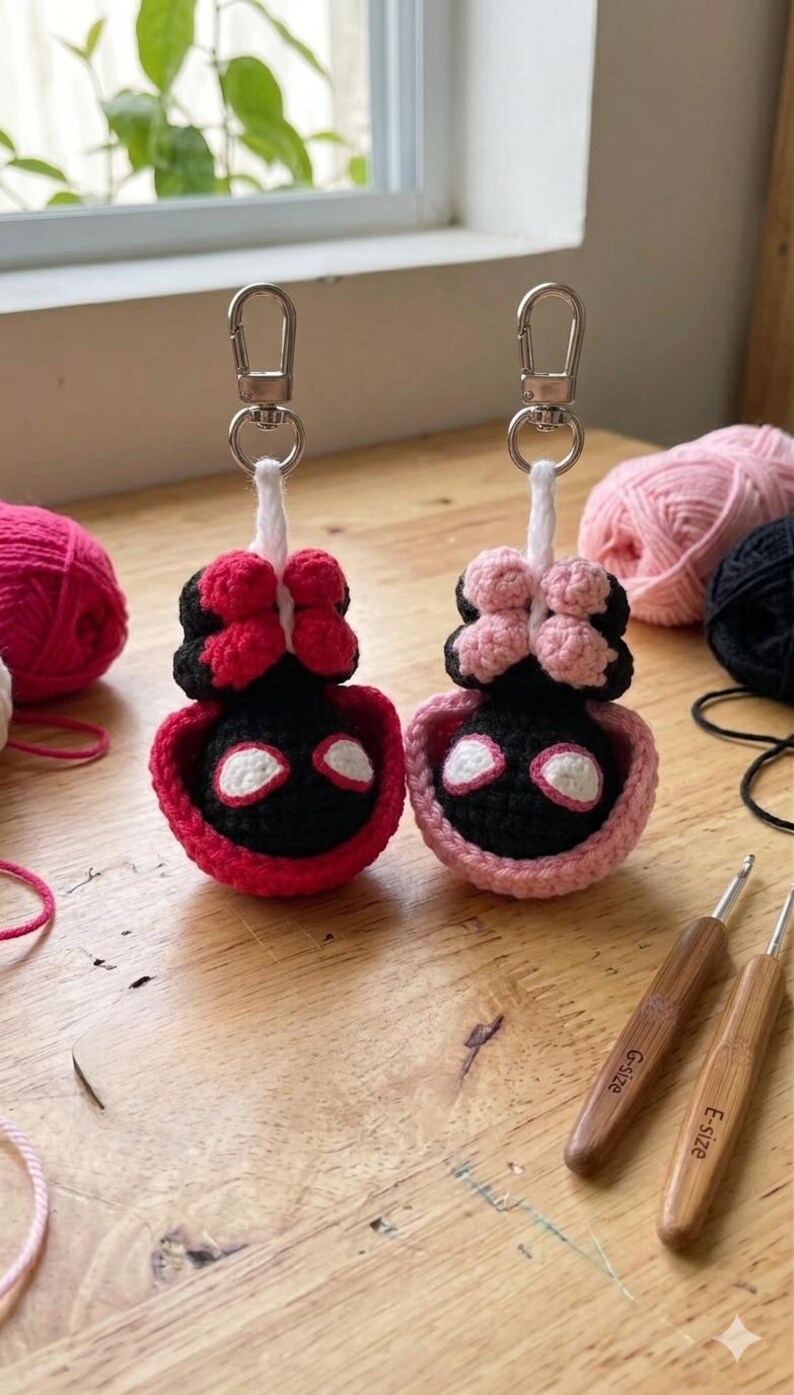 May include: Two handmade crochet keychains, one red and black, the other pink and black, each with a silver keyring. The keychains are shaped like a character with a bow. Yarn and crochet hooks are in the background.