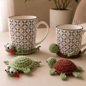 Può includere: Due tazze in ceramica bianca con un motivo geometrico a foglie nere, ciascuna appoggiata su un sottobicchiere all'uncinetto a forma di tartaruga. Un sottobicchiere è verde chiaro con una lingua rossa, l'altro è marrone. Due altri sono davanti.