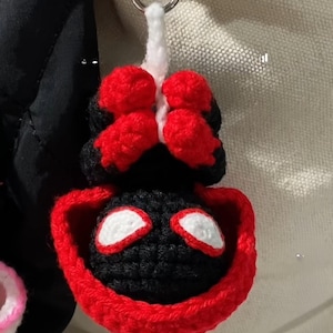 May include: Crocheted keychain featuring a black and red superhero mask design. The keychain is attached to a silver metal clasp. The mask has white eye details and a red border.