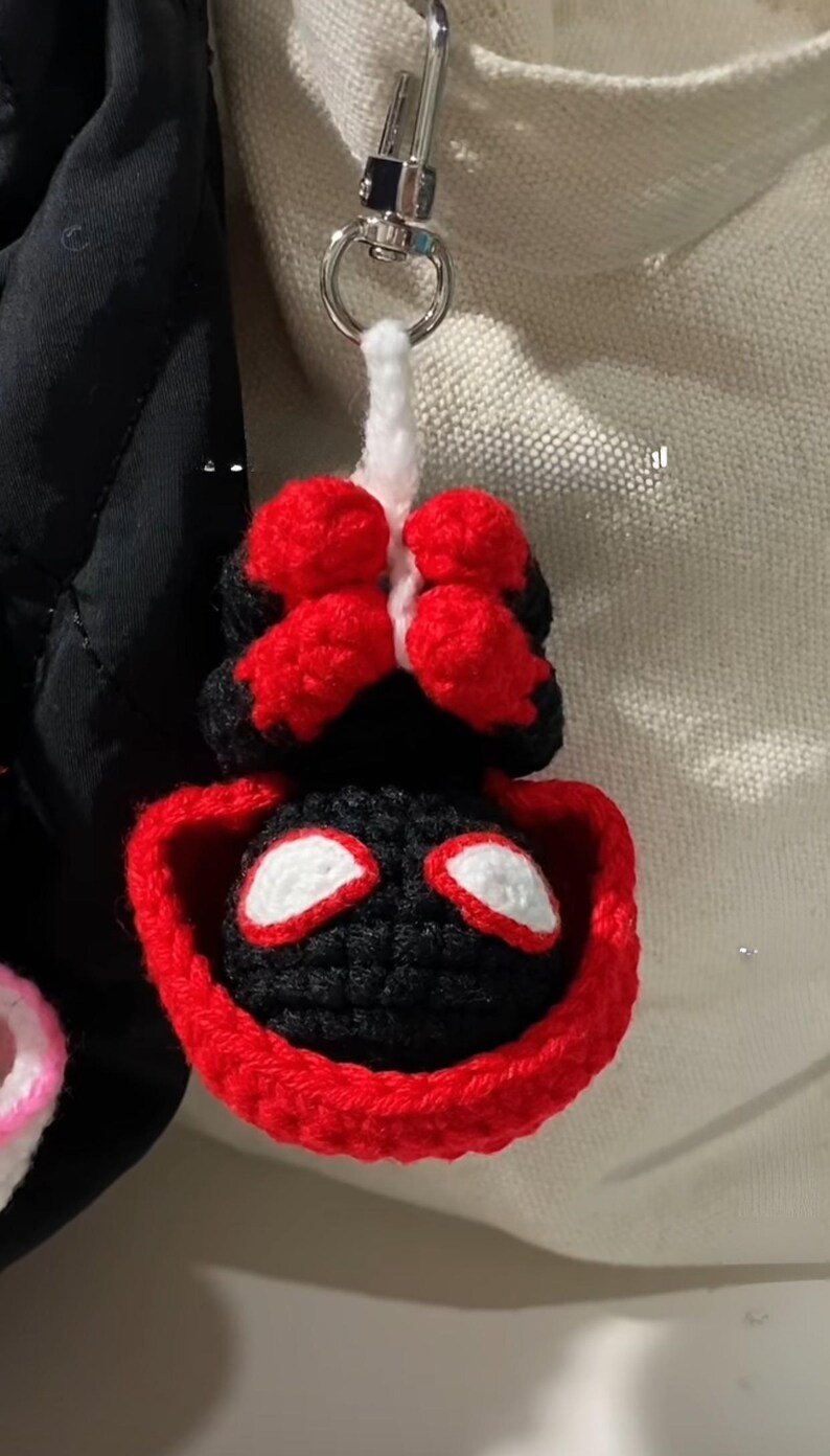 May include: Crocheted keychain featuring a black and red superhero mask design. The keychain is attached to a silver metal clasp. The mask has white eye details and a red border.