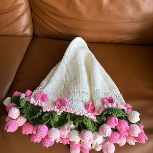 May include: A handmade, off-white crochet blanket with a scalloped edge, adorned with pink and white crochet tulips and green leaves. The blanket is folded, showcasing the floral border.
