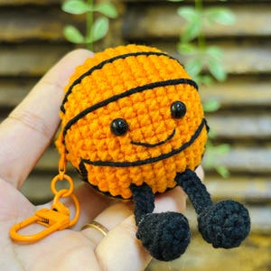 Op de afbeelding: Een handgemaakte oranje en zwarte gehaakte basketbal met een lachend gezicht, zwarte benen en een bijpassende oranje sleutelhanger. De basketbal heeft zwarte strepen en zwarte ogen. De sleutelhanger is bevestigd aan de bovenkant van de basketbal.