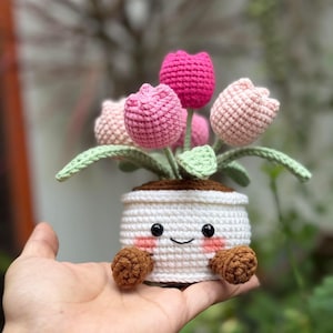 Puede incluir: Una maceta de ganchillo con una cara sonriente, que sostiene varios tulipanes rosas y rosa claro. La maceta es blanca con detalles marrones y pequeñas patas marrones. Los tulipanes tienen hojas verdes.