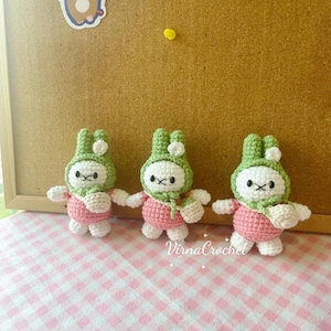 May include: Three handmade crochet bunny dolls. Each bunny has a white face, green hat, pink body, and white arms and legs. The dolls are arranged on a pink and white checkered surface. The text "VirnaCrochet" is visible.
