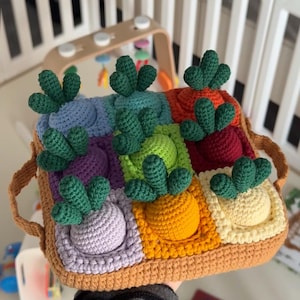 以下が含まれることがあります： 手作りの正方形デザインのかぎ針編みおもちゃ。さまざまな色の正方形があり、それぞれにかぎ針編みの野菜と緑の葉が付いています。おもちゃには茶色の縁とハンドルがあり、子供向けに設計されています。