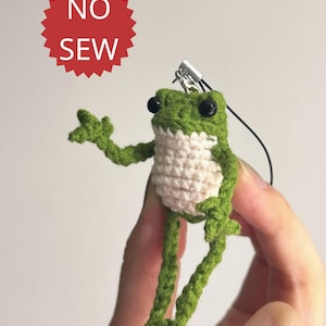 May include: A handmade crochet frog in green and white, with black eyes. The frog is holding a pose with its arms and legs extended. The image includes a red starburst with the words "NO SEW".