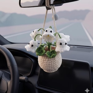 Puede incluir: Un adorno colgante de ganchillo para coche que presenta una maceta de color crema con flores blancas de lirio de los valles, hojas verdes y ojos de cuentas negras. La planta cuelga de un cordón color crema con una pequeña cuenta blanca.