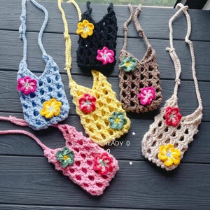 May include: Six handmade crochet crossbody bags in various colors: blue, yellow, black, brown, pink, and cream. Each bag features a floral applique in contrasting colors and a long strap. The text "READY 0" is visible.