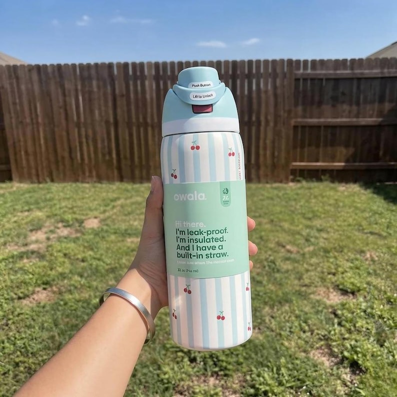 Puede incluir: Una botella de agua Owala azul claro y blanca con un patr&oacute;n de cerezas y rayas verticales. La botella tiene una pajita incorporada y el texto "Hi there, I'm leak-proof. I'm insulated. And I have a built-in straw."