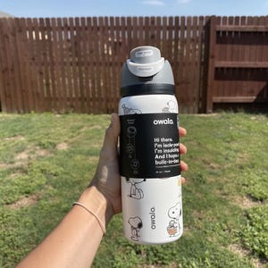 Peut inclure: Une gourde blanche Owala avec un motif Snoopy noir et blanc. La bouteille a un couvercle gris et une bande noire avec du texte. La bouteille est tenue devant une clôture en bois et de l'herbe verte.