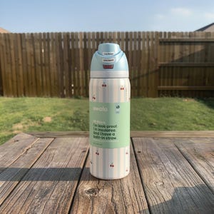 Puede incluir: Una botella de agua Owala blanca y azul claro con un patr&oacute;n de cerezas y una tapa azul claro. La botella tiene una banda verde con el texto "Hi there. I'm leak-proof. I'm insulated. And I have a built-in straw."
