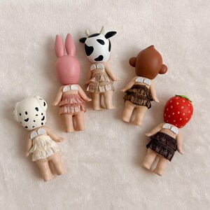 Puede incluir: Cinco peque&ntilde;as figuras con cabezas de animales: un d&aacute;lmata, un conejito rosa, una vaca, un mono y una fresa. Cada figura tiene una peque&ntilde;a falda con volantes y una parte superior blanca. Las figuras est&aacute;n dispuestas sobre una superficie blanca y suave.