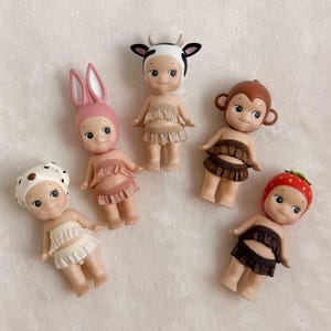 Puede incluir: Cinco peque&ntilde;as figuras coleccionables con sombreros tem&aacute;ticos de animales y faldas con volantes. Las figuras incluyen una vaca, un conejito, un mono, una fresa y un sombrero con lunares. Cada figura tiene una cara sonriente.