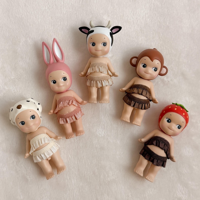 Puede incluir: Cinco peque&ntilde;as figuras coleccionables con sombreros tem&aacute;ticos de animales y faldas con volantes. Las figuras incluyen una vaca, un conejito, un mono, una fresa y un sombrero con lunares. Cada figura tiene una cara sonriente.