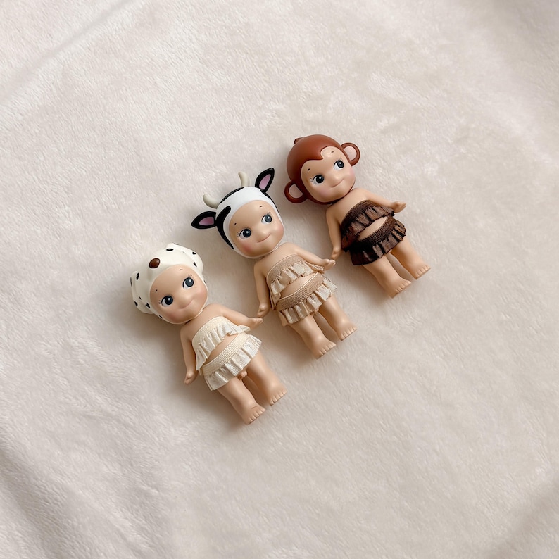 Puede incluir: Tres peque&ntilde;as figuras decorativas. Una es una vaca, otra es un mono y otra es un d&aacute;lmata. La figura de la vaca tiene un sombrero con estampado de vaca en blanco y negro con cuernos negros. La figura del mono tiene una cabeza marr&oacute;n y una falda marr&oacute;n. La figura del d&aacute;lmata tiene un sombrero blanco con manchas marrones.