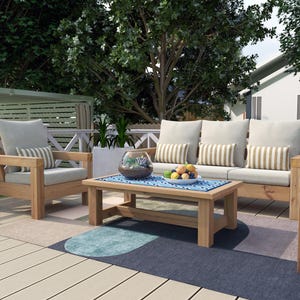 May include: Outdoor wooden furniture set with light beige cushions and striped pillows. The set includes a loveseat, two armchairs, and a coffee table. A blue and white patterned rug and a bowl of fruit are on the table.