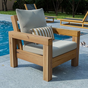 Può includere: Una poltrona da esterno in legno con cuscino e schienale beige chiaro. Un cuscino più piccolo a righe marroni e bianche poggia sulla sedia. La sedia è in legno chiaro ed è posizionata vicino a una piscina.