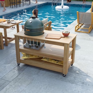 May include: A wooden outdoor grilling station with a green egg-shaped grill on top. The station has a lower shelf with bags of charcoal and a top shelf with a cutting board and bowls. Utensils hang below the top shelf. The station is on wheels.