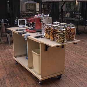Puede incluir: Un carrito de caf&eacute; m&oacute;vil con una m&aacute;quina de espresso roja, un molinillo de caf&eacute; y frascos de vidrio llenos de galletas. El carrito es de madera clara y tiene un cubo de basura y botellas de agua en su interior. El carrito est&aacute; sobre ruedas y est&aacute; instalado al aire libre.