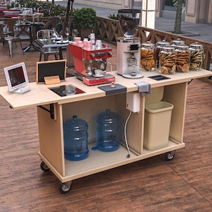 Puede incluir: Una estaci&oacute;n de caf&eacute; m&oacute;vil con una m&aacute;quina de espresso roja, un molinillo de caf&eacute; y frascos de vidrio llenos de galletas. La estaci&oacute;n incluye una peque&ntilde;a tableta, una pizarra y un cubo de basura. Dos botellas de agua grandes se almacenan debajo.