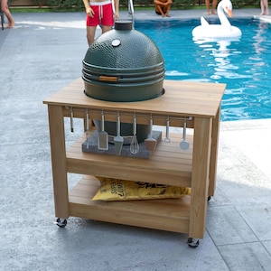 May include: A green ceramic grill sits on a wooden cart with a lower shelf and a set of grilling tools. The cart has wheels for easy movement. The grill is near a swimming pool.