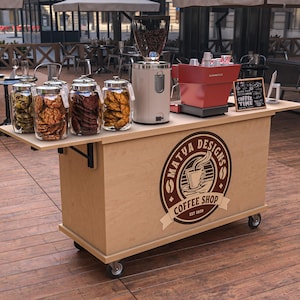 Puede incluir: Un carrito de cafeter&iacute;a m&oacute;vil con acabado de madera clara. Cuenta con un molinillo de caf&eacute;, una m&aacute;quina de espresso y frascos de galletas. El carrito tiene el logotipo "MATVA DESIGNS COFFEE SHOP" y tiene ruedas.