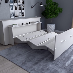May include: A white, modern, fold-out bed with a built-in storage unit. The bed is partially extended, revealing a multi-panel design. The storage unit has a shelf with books and decorative items. The room has a grey rug and a potted plant.