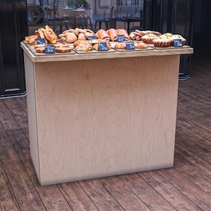 May include: A wooden bakery display case filled with various baked goods, including pastries, breads, and tarts. Small chalkboard signs with numbers are placed in front of the baked goods. The display case is on a wooden floor.