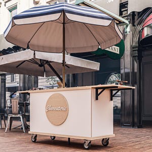 Op de afbeelding: Een witte mobiele charcuteriekar met een gestreepte parasol. De kar heeft een houten aanrecht en een rond bord met de tekst "Charcuterie". De kar staat op wielen en heeft een houten vloer.