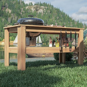 May include: A wooden outdoor grilling station with a black grill and a stainless steel firebox. The station has a lower shelf and hooks for utensils. Various grilling tools and condiments are visible. The background features a lake and trees.