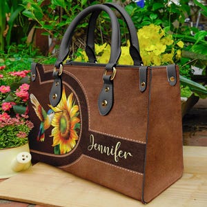 May include: Brown handbag with a hummingbird and sunflower design. The bag features a dark brown handle and accents, with the name "Jennifer" in gold script. The bag is made of a suede-like material.