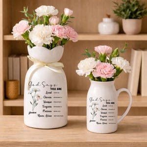 May include: Two white ceramic vases with floral arrangements. One vase is a unique shape with a bow, the other is a mug. Both have text that reads "God Says You Are Loved, Chosen, Unique, Forgiven, Precious, Never Alone."