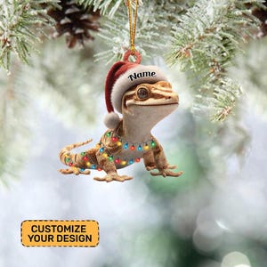 May include: A Christmas ornament featuring a gecko wearing a Santa hat and a string of colorful lights. The ornament has the word "Name" on the hat and the text "Customize Your Design" on a yellow rectangle.