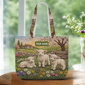 May include: A personalized tote bag featuring a pastoral scene with three lambs in a field of wildflowers. The bag has a white handle and the text "YOUR NAME" on a green label. The background shows a house, trees, and a stone fence.