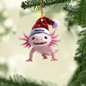 May include: A pink axolotl Christmas ornament wearing a red Santa hat with white trim and snowflakes. The hat has the word "Name" on it. The ornament is hanging from a gold string, with green pine branches in the background.