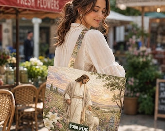 Puede incluir: Un bolso tote beige con una escena pastoral que representa una figura con una túnica y un cordero, sobre un fondo de campo y cielo. El bolso tiene una correa a juego y el texto "YOUR NAME" en un letrero verde.