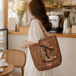May include: A brown tote bag featuring an embossed western-style design of a cowboy hat and a pair of cowboy boots. The bag has a rectangular shape with a shoulder strap and includes a small rectangular label with the word Name printed on it.