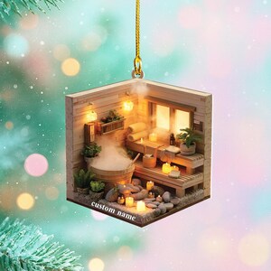 May include: A miniature sauna ornament with a wooden exterior, featuring a sauna interior with a wooden tub, benches, and lit candles. The ornament has the text "custom name" on the bottom and is suspended by a gold string.