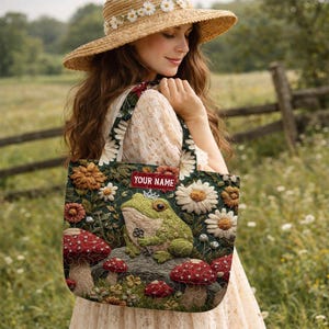 May include: A green and floral tote bag featuring a frog wearing a crown, mushrooms, and the text "YOUR NAME" on a red label. The bag has a dark green background with white and yellow flowers.
