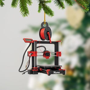 May include: A miniature 3D printer ornament in black and red, with a spool of red filament. The ornament is suspended from a gold string and a black loop. The word "Name" is printed on the base of the printer.