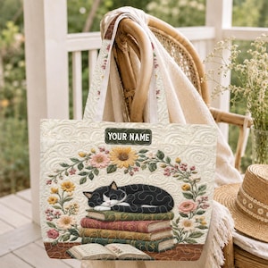 May include: Cream-colored tote bag with a quilted design. The bag features a black and white cat sleeping on a stack of books, surrounded by a floral wreath. The text "YOUR NAME" is displayed on a black nameplate.