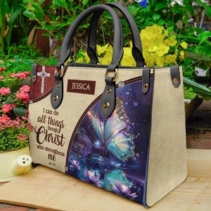 May include: Beige handbag with black handles and gold-colored hardware. The bag features a butterfly design and the text "I can do all things through Christ who strengthens me." A brown patch displays the name "JESSICA".