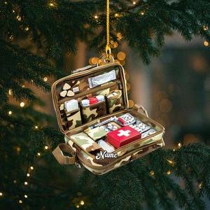 May include: A camouflage-patterned first aid kit ornament, open to display medical supplies. The kit includes a red box with a white cross, pills, a syringe, and other items. The word "Name" is printed on the front.