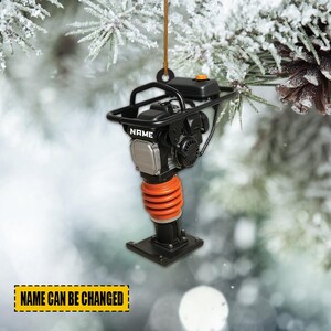 May include: A black and orange construction equipment ornament with the word "NAME" printed on the side. The ornament has a handle and an orange spring. The text "NAME CAN BE CHANGED" is printed on a yellow rectangle.
